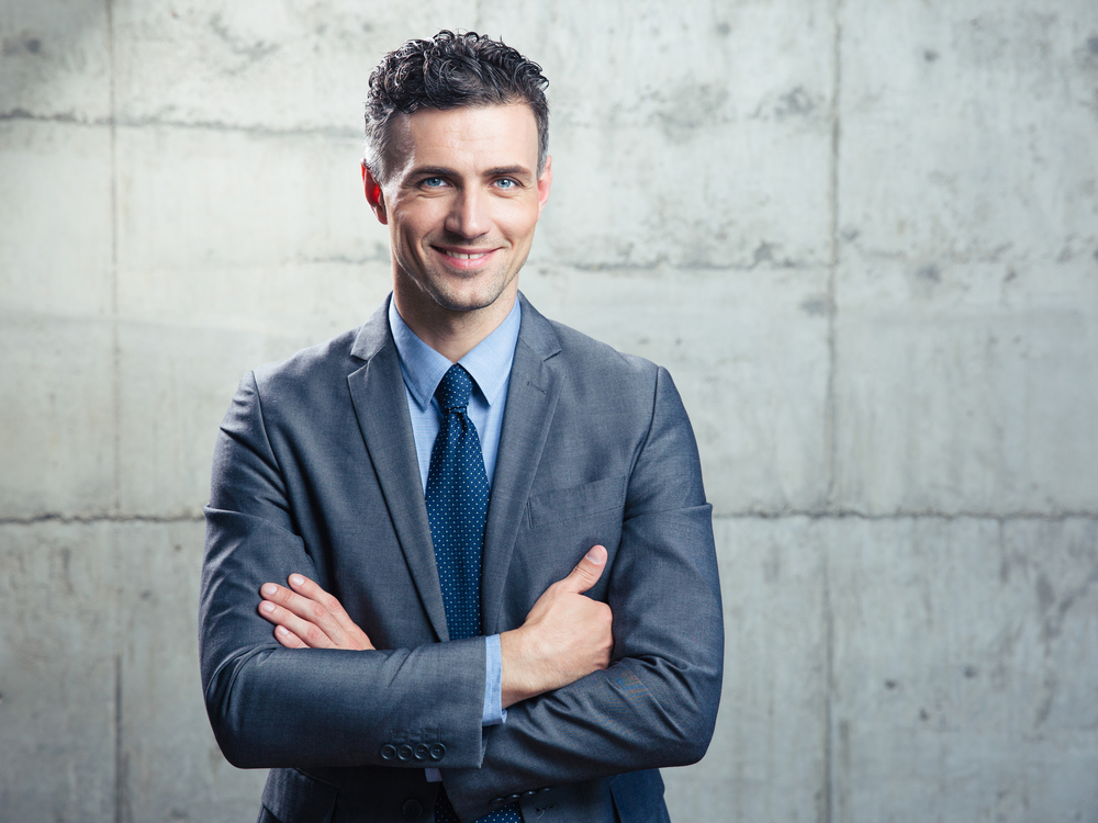 Portrait of a smiling handsome businessman with arms folded over concrete wall